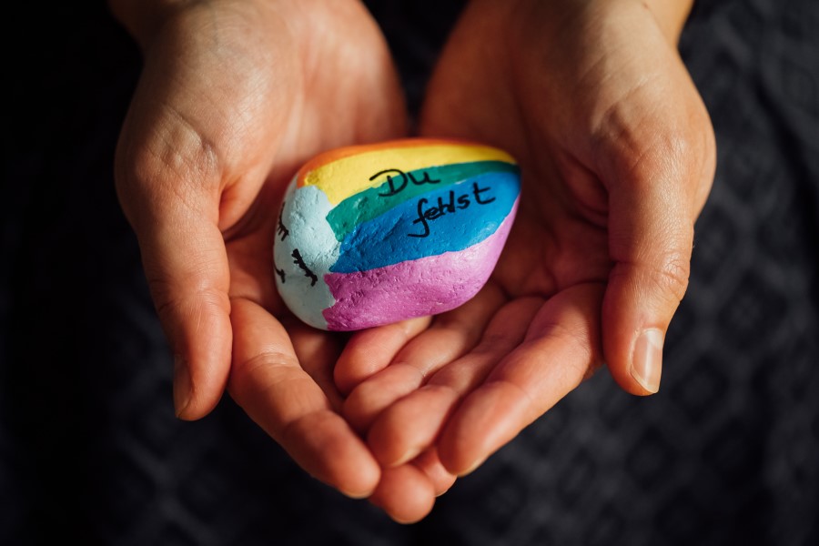 Zwei Hände halten einen Regenbogen-Stein mit der Aufschrift "Du fehlst"